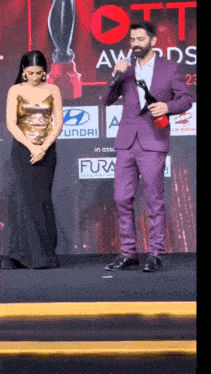 a man in a purple suit is holding a trophy and speaking into a microphone while a woman looks on