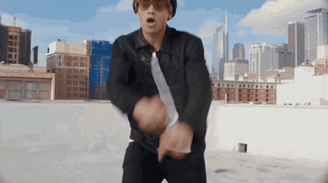 a man wearing sunglasses and a black jacket is dancing on a rooftop in front of a city skyline
