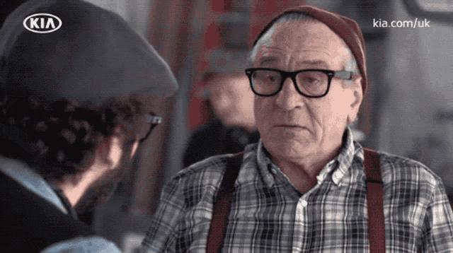 a man talking to an older man with glasses and suspenders and a kia logo on his hat