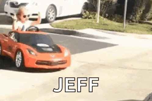 a little boy is driving a red toy car on a street .