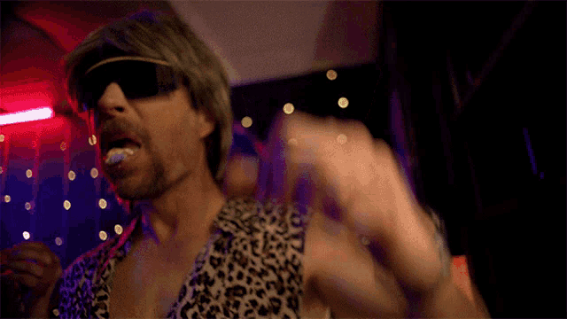 a man wearing sunglasses and a leopard print shirt is dancing in a dark room