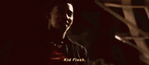 a man in a black jacket is talking to another man in a dark room and says `` kid flash '' .