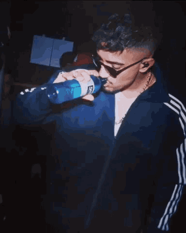 a man wearing sunglasses and a blue jacket is drinking from a sprite bottle