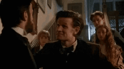 a man in a tuxedo and bow tie is talking to another man in a room .