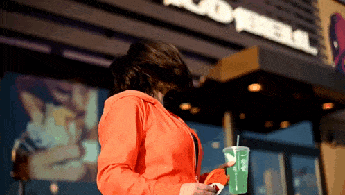 a woman in an orange jacket holds a drink in front of a taco bell restaurant