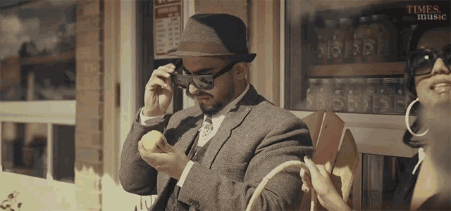 a man wearing a hat and sunglasses sits next to a woman wearing hoop earrings and a sign that says times of music