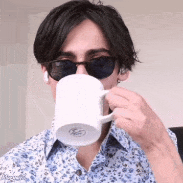 a young man wearing sunglasses and a floral shirt is drinking from a white mug .