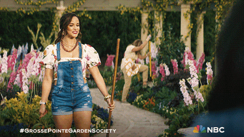 a woman in overalls is holding a broom in a garden with a nbc logo behind her