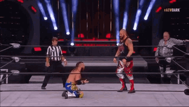 a wrestler is kneeling down in a wrestling ring while another wrestler stands in the corner .