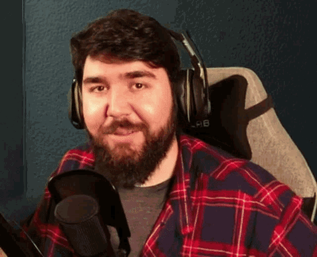 a man with a beard is wearing headphones and a plaid shirt while sitting in front of a microphone .