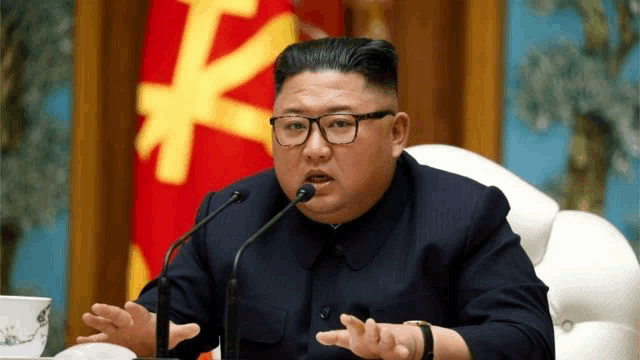 a man wearing glasses is talking into a microphone in front of a flag with a k on it