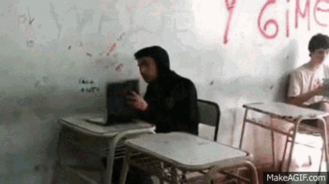 a man in a hoodie is sitting at a desk in a classroom using a laptop computer .