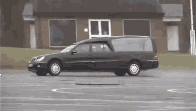 a black funeral car is driving in a parking lot in front of a building .