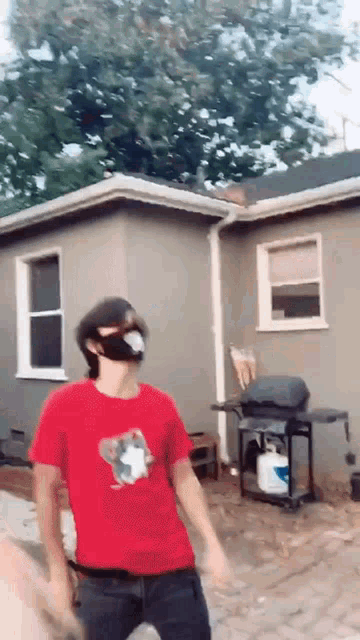 a man wearing a red t-shirt and a mask is standing in front of a house .