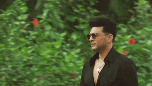 a man in a black shirt and sunglasses stands in front of a bush