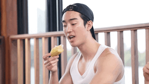 a man wearing a baseball cap and a white tank top is eating corn on the cob