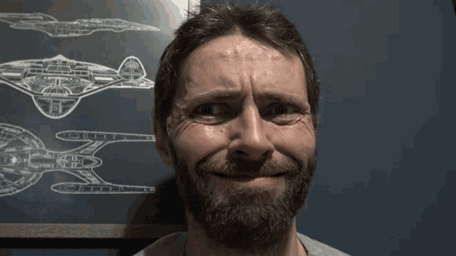 a man with a beard is smiling in front of a drawing of a spaceship