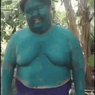 a fat man with blue paint on his body is standing in the woods and smiling .