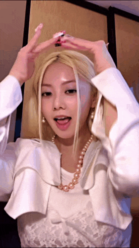 a woman in a white shirt and pearls is making a heart shape with her hands .