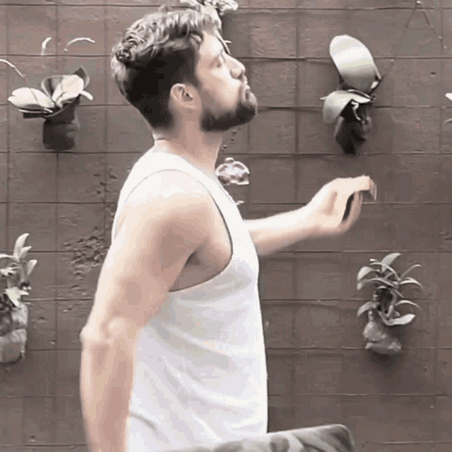 a man in a white tank top is standing in front of a wall with plants hanging from it