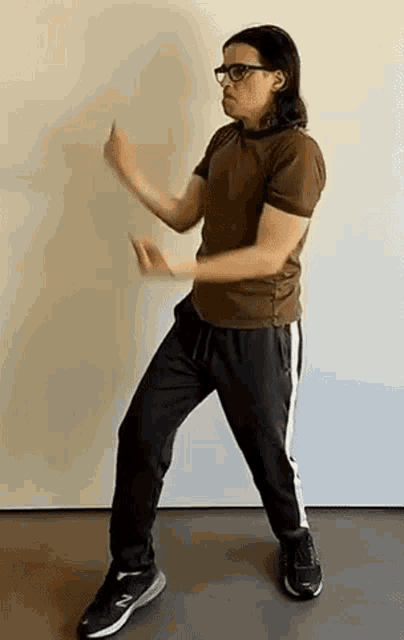 a man in a brown shirt and black pants is standing in front of a white wall .
