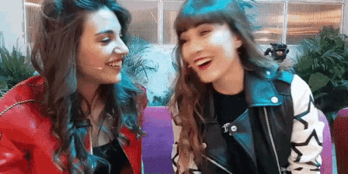 two women are laughing and looking at each other while sitting next to each other .