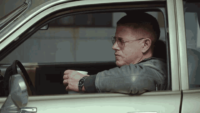 a man wearing glasses sits in a car holding a cup of coffee