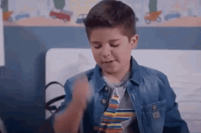 a young boy is sitting on a bed wearing a denim jacket and tie .