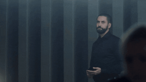 a man with a beard wearing a black shirt stands in a dark room