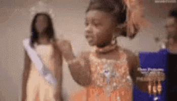 a little girl in an orange dress is standing next to a woman in a crown .