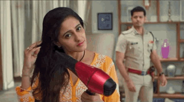 a woman is blow drying her hair in a living room while a man in a police uniform stands behind her .