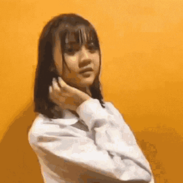 a young girl in a white shirt is standing in front of a yellow wall and touching her hair .