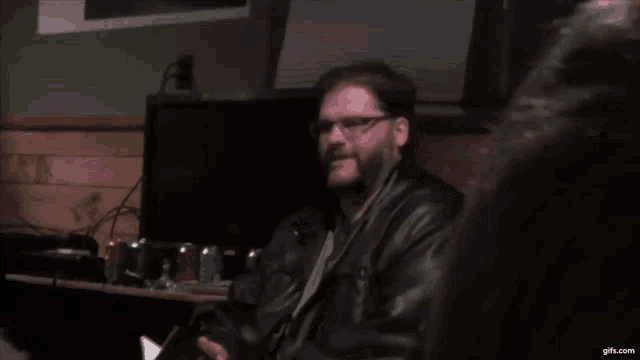a man wearing glasses and a black leather jacket is sitting in front of a computer monitor