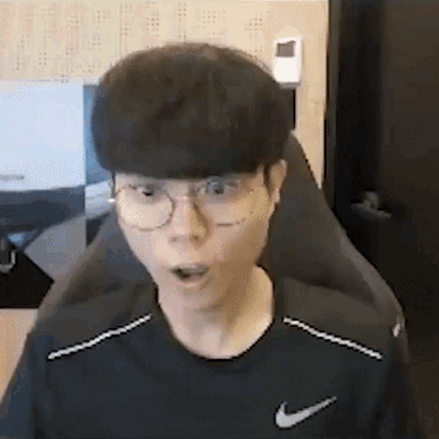a young man wearing glasses and a nike shirt is sitting in a chair and making a surprised face .