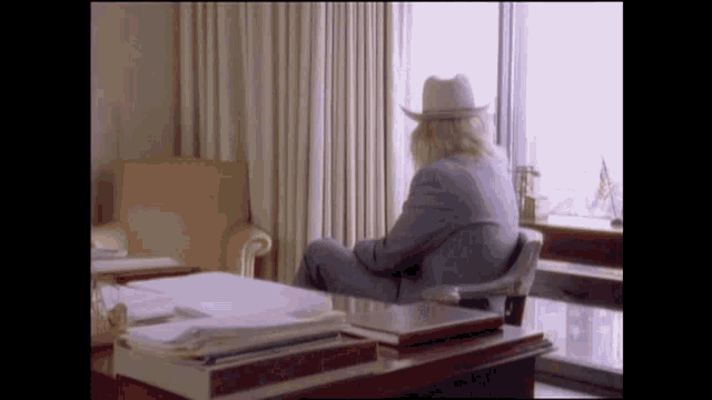 a man in a cowboy hat sits at a desk in front of a large window
