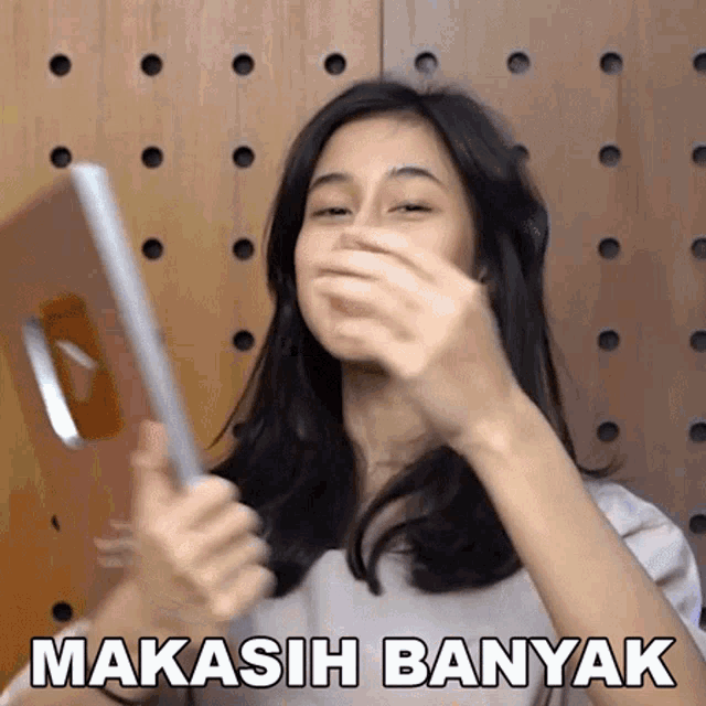 a woman covering her mouth while holding a tablet with the words " makasih banyak " written below her