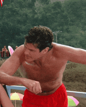 a shirtless man in red shorts stands on a beach