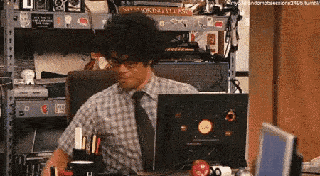 a man in a plaid shirt and tie is sitting at a desk in front of a computer screen .