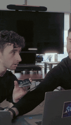 a man holding a cell phone talks to another man in front of a laptop with a sticker that says sc