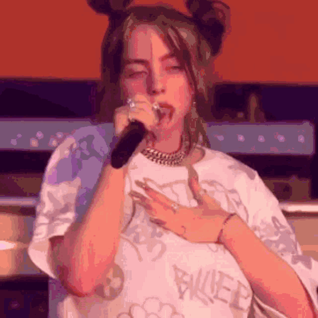 billie eilish is singing into a microphone while wearing a white shirt with the word billie eilish on it .