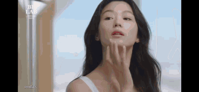 a woman in a white tank top is applying a cream to her face .
