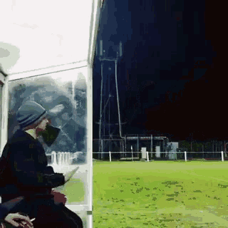 a man in a beanie looks out a window at a soccer field at night