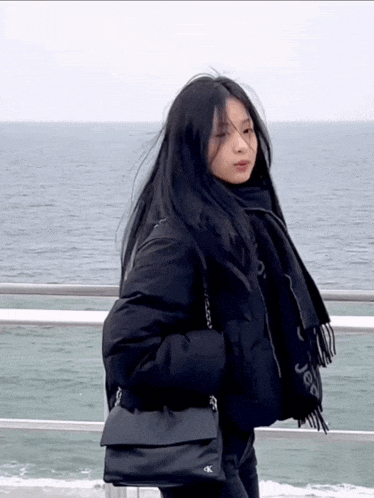 a woman wearing a black jacket and scarf is standing in front of the ocean