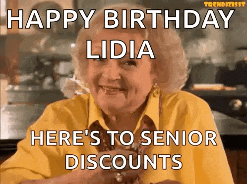an elderly woman is sitting at a table with a glass of wine and a happy birthday greeting .