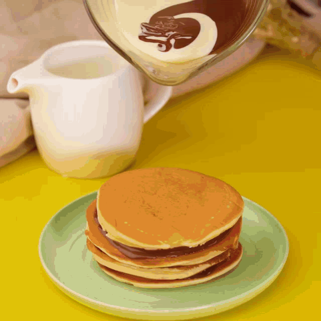 a stack of pancakes on a green plate with chocolate sauce being poured on them