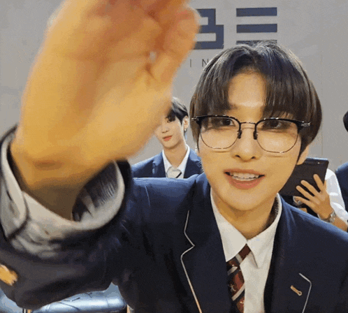 a young man wearing glasses and a suit waves his hand