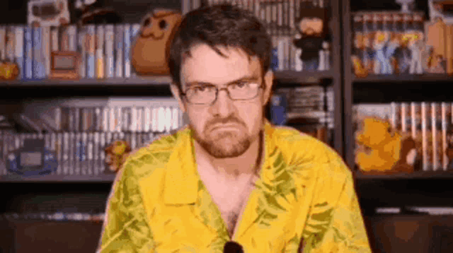 a man wearing glasses and a yellow shirt is sitting in front of a shelf filled with video games .