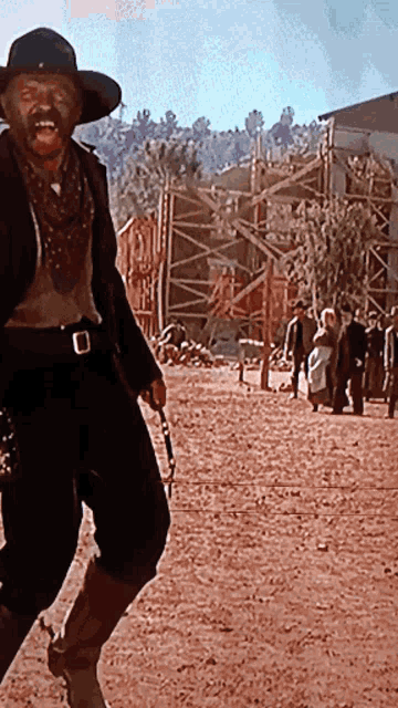 a man in a cowboy hat stands in a dirt field holding a gun