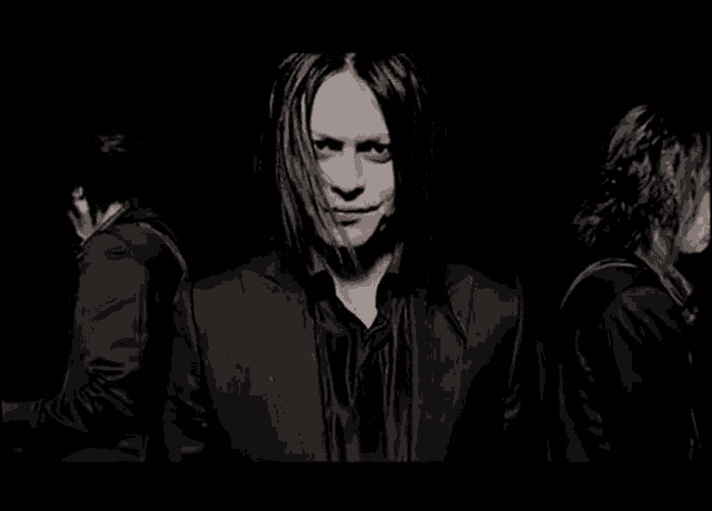 a man with long black hair is standing next to two other men in a black suit .