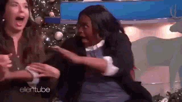 a woman is sitting in front of a christmas tree and laughing while another woman looks on .